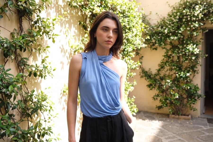 Woman wearing a cornflower blue drape high-neck top in a sunlit courtyard