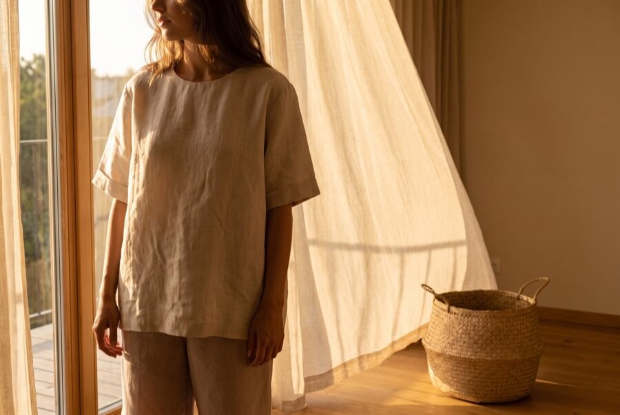 Woman wearing an oversized natural linen top beside a billowing curtain in golden light