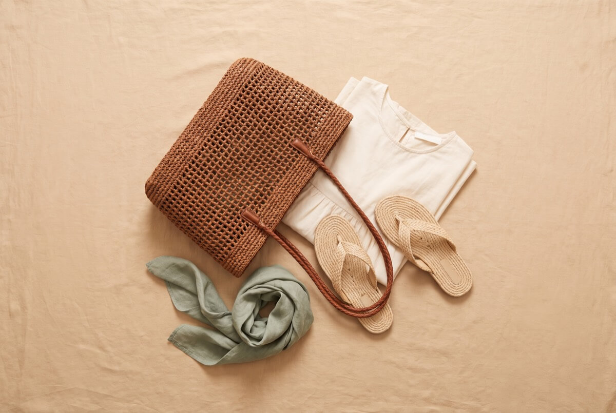 Structured brown raffia tote with an open weave and a woven leather strap, laid flat with a cream dress, woven sandals and a sage scarf