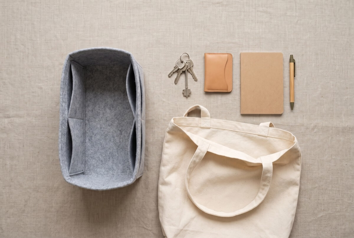 Soft grey bag organiser insert laid flat beside an empty cream canvas tote, with keys, a tan card holder, a notebook and a pen