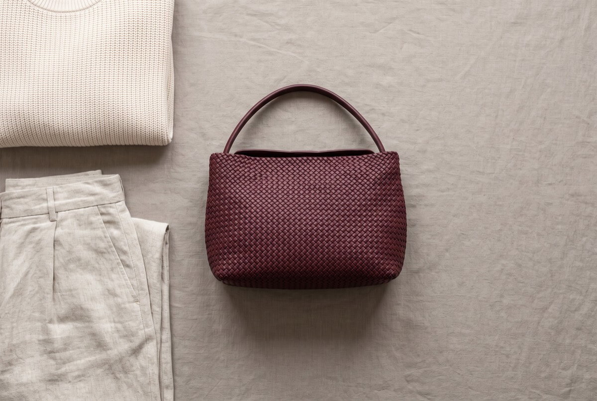 Burgundy handwoven handbag with a leather top handle, laid flat beside a cream knit jumper and folded oatmeal trousers