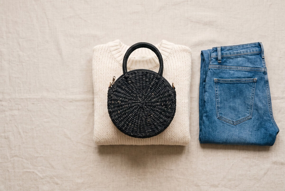 Small round black handwoven palm purse with a top handle and zip, resting on a folded cream knit jumper beside blue jeans