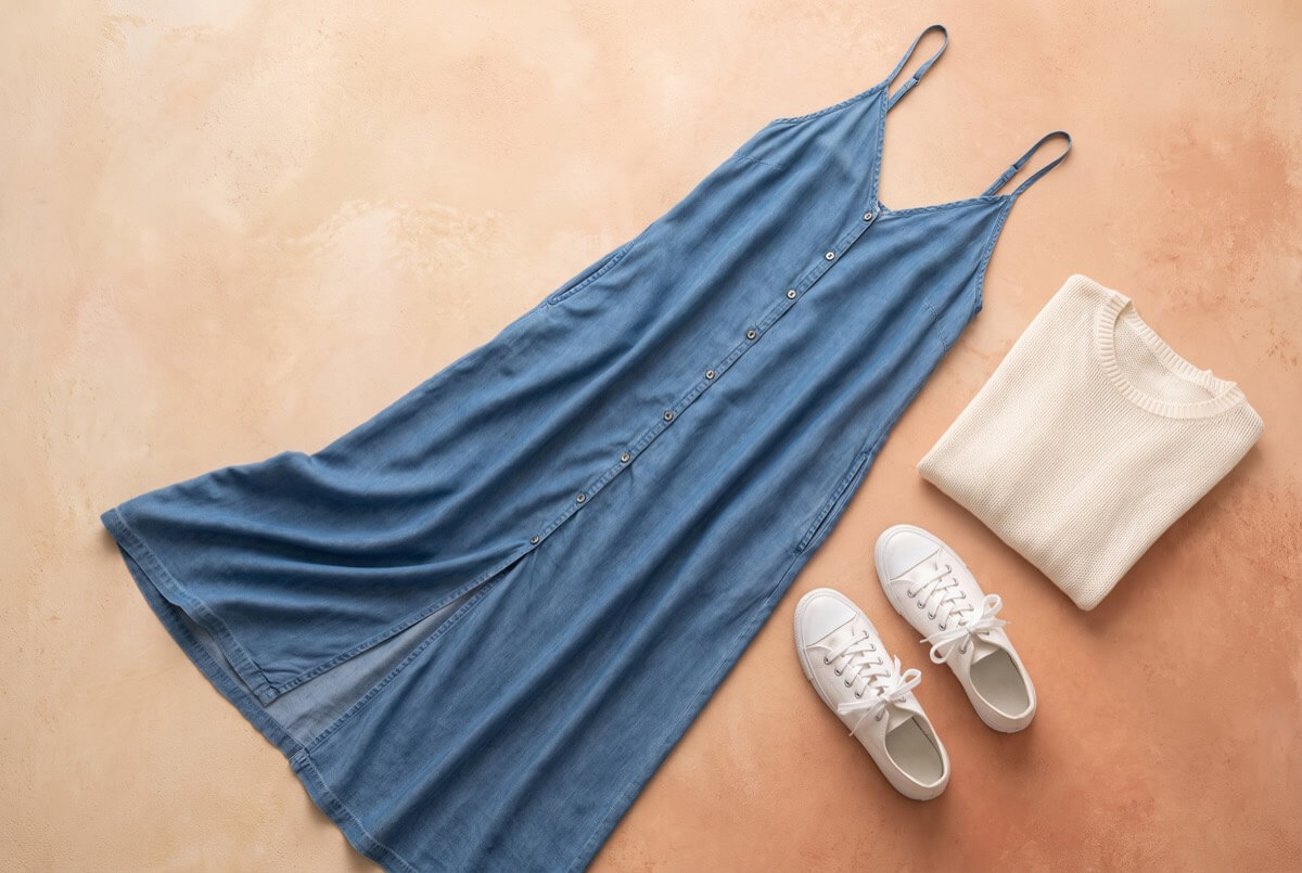 Denim-blue midi slip dress with front buttons laid flat beside white sneakers, sustainable fashion