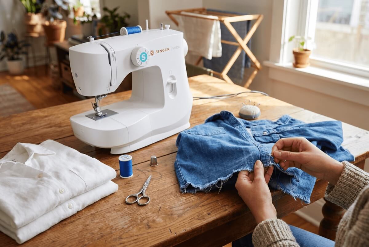 Hands mending a denim piece beside a sewing machine and fabric scissors on a wooden table - beginner upcycling fixes for a zero waste wardrobe