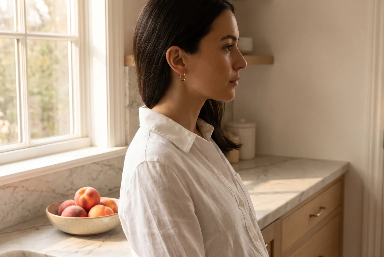 Woman in profile by a bright kitchen window wearing small polished gold hoop earrings and an open white linen shirt