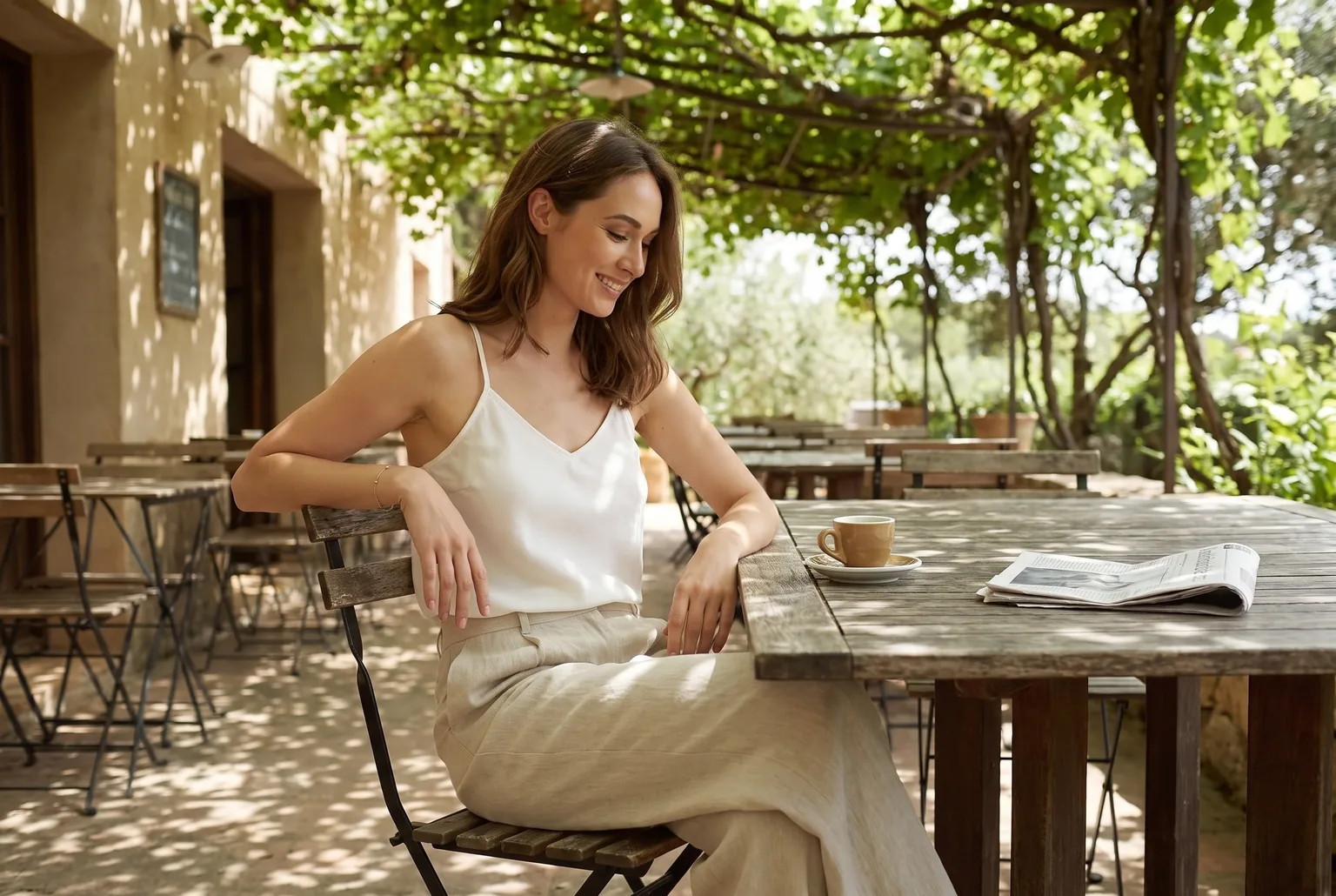 Woman at a shaded cafe table wearing a cream bias-cut camisole tucked into oat linen pants