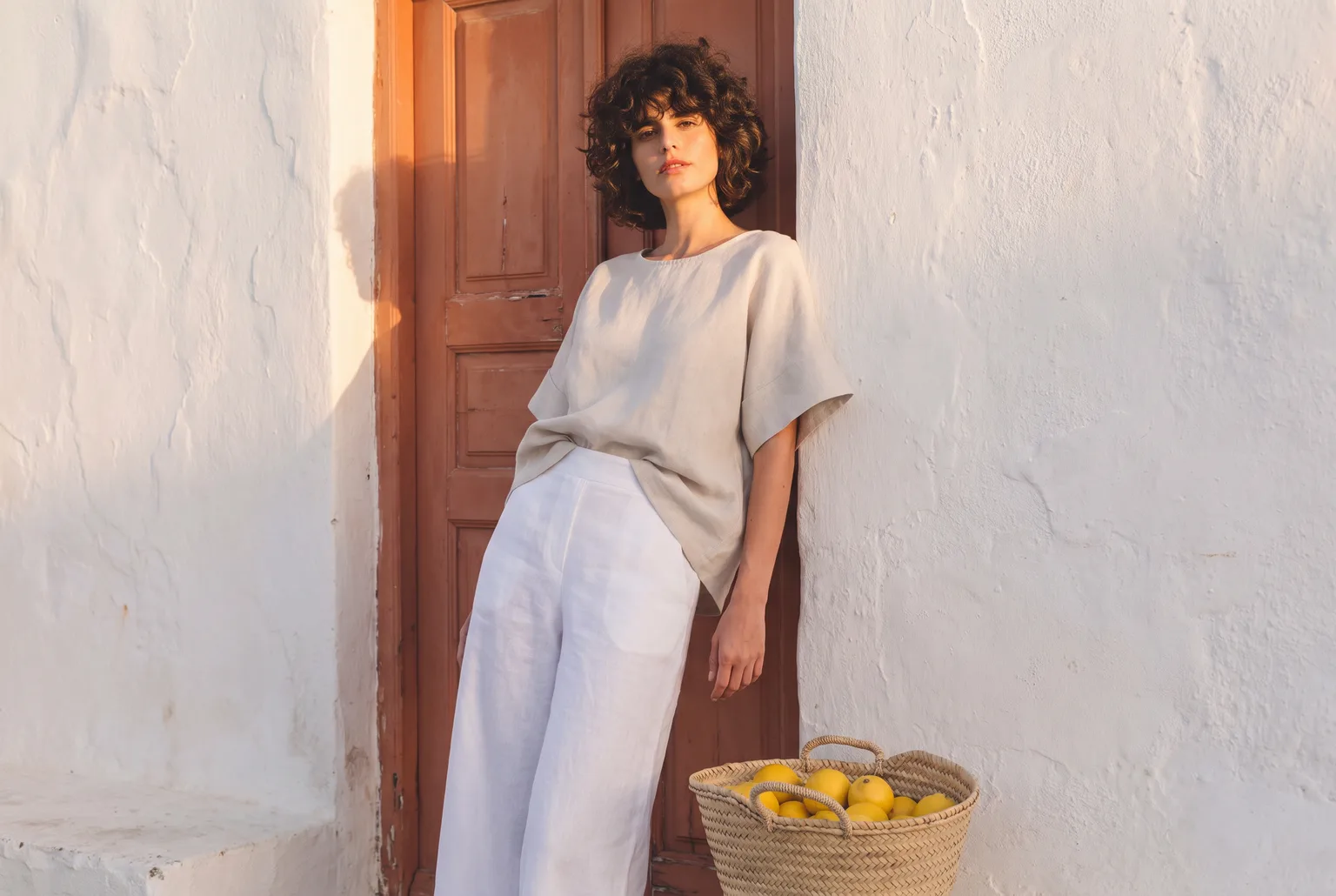 Woman in a terracotta doorway wearing a cream oversized linen top half-tucked into white linen pants